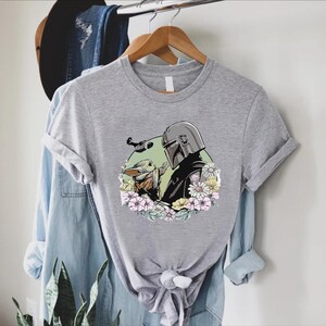 Peut inclure: Un t-shirt gris chin&eacute; avec un motif d'un personnage avec un casque et une petite cr&eacute;ature, entour&eacute;s de fleurs. Le motif est plac&eacute; sur un cercle vert. Le t-shirt est suspendu &agrave; un cintre en bois.