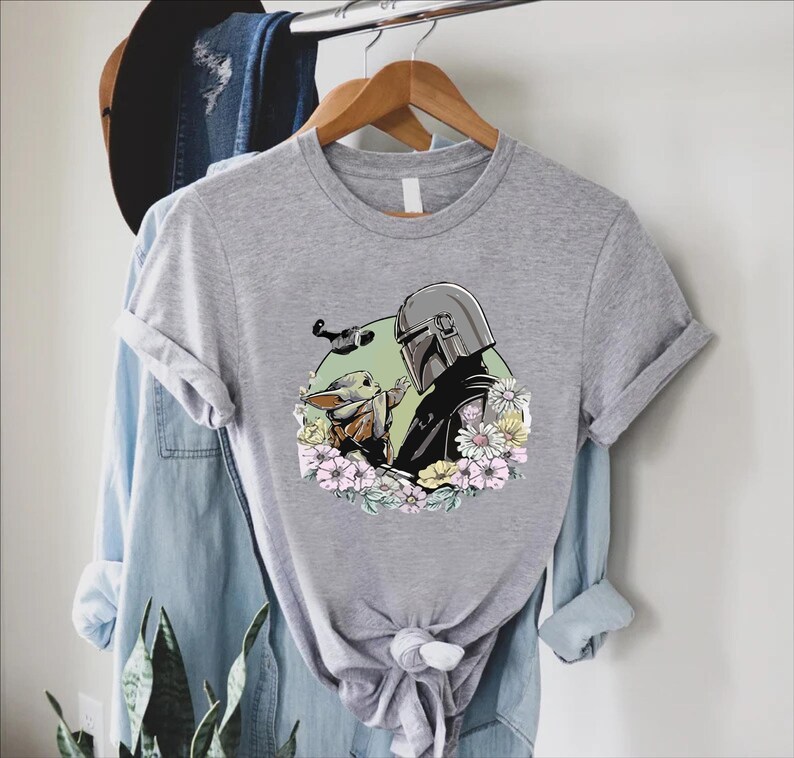Peut inclure: Un t-shirt gris chin&eacute; avec un motif d'un personnage avec un casque et une petite cr&eacute;ature, entour&eacute;s de fleurs. Le motif est plac&eacute; sur un cercle vert. Le t-shirt est suspendu &agrave; un cintre en bois.