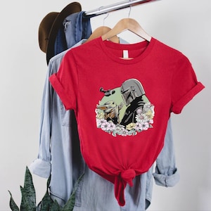 Peut inclure: T-shirt rouge avec un motif d'un personnage avec un casque et une petite cr&eacute;ature, entour&eacute;s de fleurs. Le motif est centr&eacute; sur le t-shirt. Le t-shirt est suspendu &agrave; un portant avec d'autres v&ecirc;tements.
