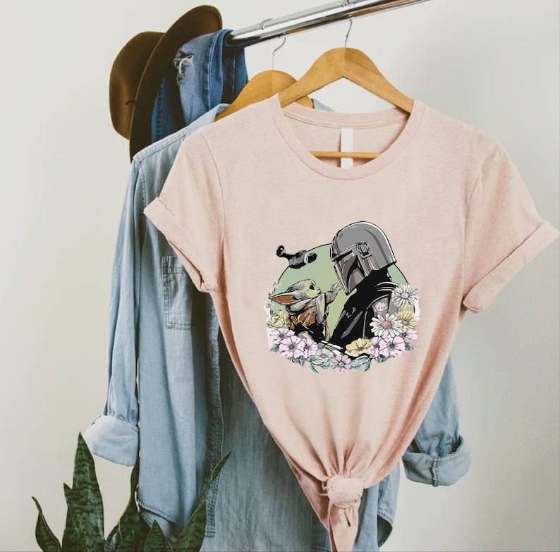 Peut inclure: Un t-shirt rose clair avec un motif d'un personnage portant un casque et une petite cr&eacute;ature, entour&eacute;s de fleurs. Le t-shirt est suspendu &agrave; un cintre en bois avec une chemise en jean bleue et un chapeau marron en arri&egrave;re-plan.