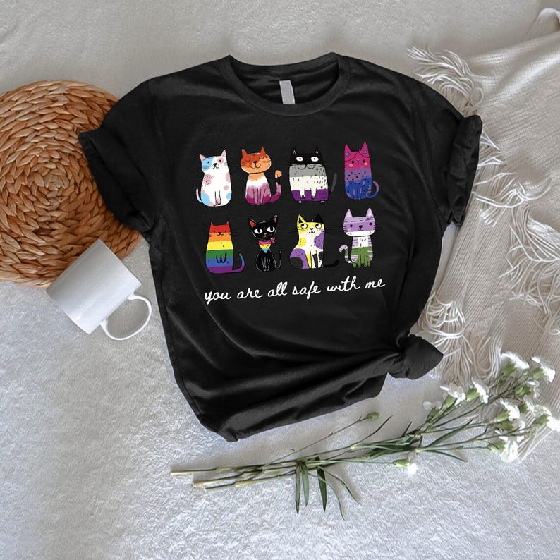 May include: Black t-shirt featuring a colorful graphic of nine stylized cats, each with a unique pattern and color scheme. The text "you are all safe with me" is printed below the cats. The shirt is made of a soft material.