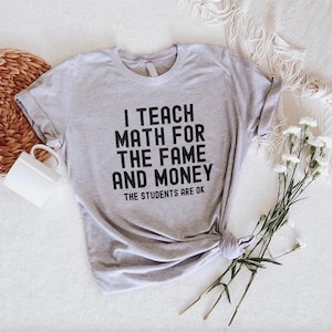May include: A light gray t-shirt with the text "I TEACH MATH FOR THE FAME AND MONEY. THE STUDENTS ARE OK" in black. The shirt is knotted at the bottom and is next to a white mug and flowers.