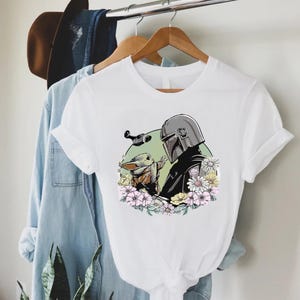 Peut inclure: T-shirt blanc avec un motif de The Mandalorian et Grogu entour&eacute;s de fleurs. Le motif comprend une figurine casqu&eacute;e et un petit personnage &agrave; la peau verte. Le t-shirt est suspendu &agrave; un cintre en bois avec d'autres v&ecirc;tements.