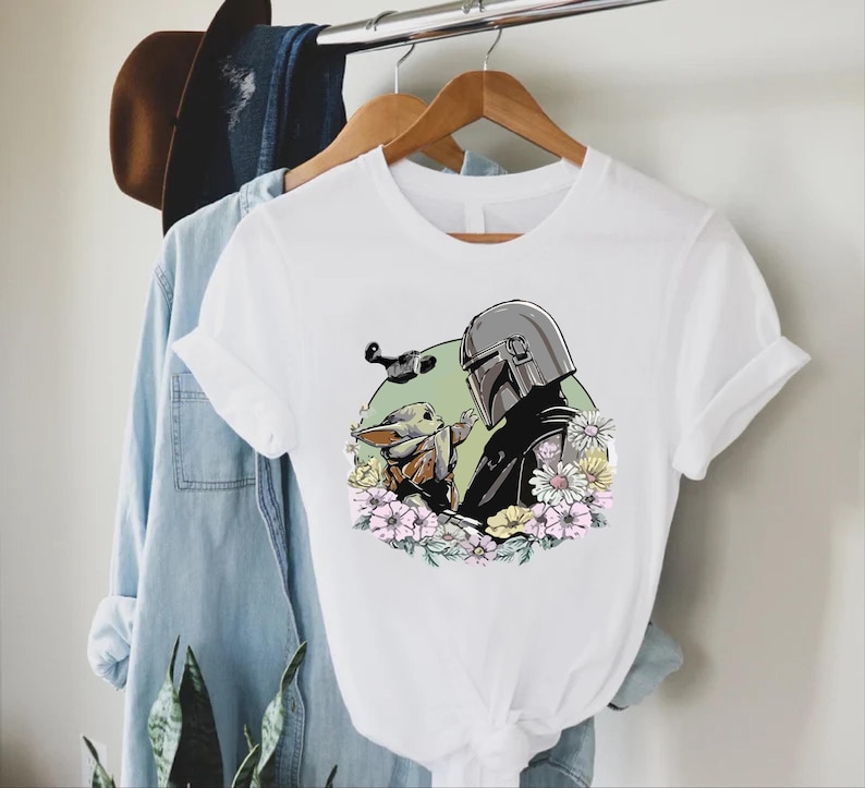 Peut inclure: T-shirt blanc avec un motif de The Mandalorian et Grogu entour&eacute;s de fleurs. Le motif comprend une figurine casqu&eacute;e et un petit personnage &agrave; la peau verte. Le t-shirt est suspendu &agrave; un cintre en bois avec d'autres v&ecirc;tements.