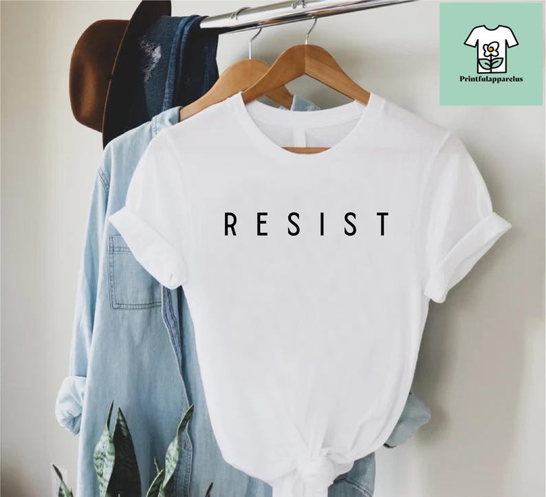 以下が含まれることがあります： 前面に黒で「RESIST」とプリントされた白いTシャツです。