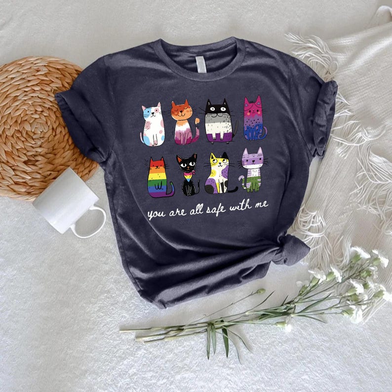May include: Navy blue t-shirt featuring a colorful illustration of nine cartoon cats with various patterns and colors. The text "you are all safe with me" is printed below the cats. The shirt is made of soft material.