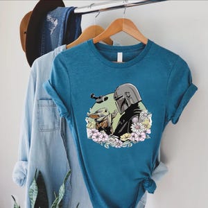 Peut inclure: T-shirt bleu sarcelle avec un motif d'un personnage avec un casque et une petite cr&eacute;ature, entour&eacute;s de fleurs. Le motif est sur un t-shirt &agrave; manches courtes suspendu &agrave; un cintre en bois. Une chemise en jean et un chapeau sont &eacute;galement visibles.
