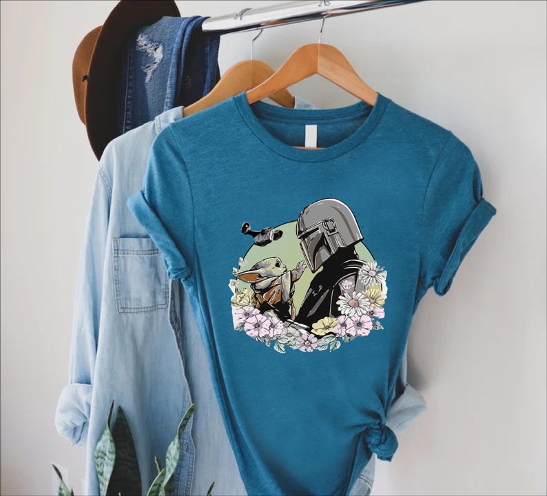 Peut inclure: T-shirt bleu sarcelle avec un motif d'un personnage avec un casque et une petite cr&eacute;ature, entour&eacute;s de fleurs. Le motif est sur un t-shirt &agrave; manches courtes suspendu &agrave; un cintre en bois. Une chemise en jean et un chapeau sont &eacute;galement visibles.