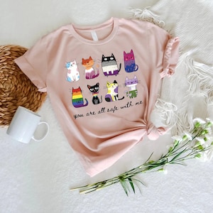 May include: A light pink t-shirt featuring a colorful graphic of nine cartoon cats with various patterns and colors. The text "you are all safe with me" is printed below the cats. The shirt is displayed on a white surface with a woven basket, a white mug, and flowers.