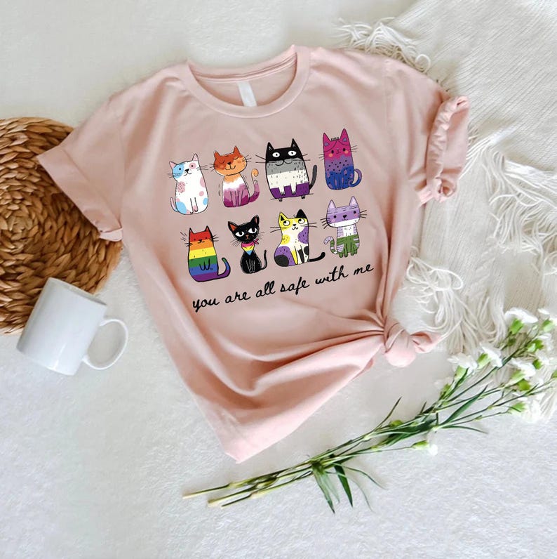 May include: A light pink t-shirt featuring a colorful graphic of nine cartoon cats with various patterns and colors. The text "you are all safe with me" is printed below the cats. The shirt is displayed on a white surface with a woven basket, a white mug, and flowers.