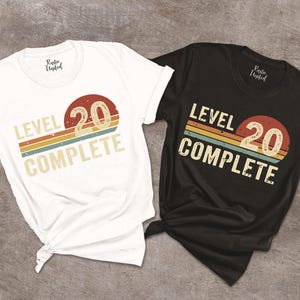 May include: Two t-shirts, one white and one black, with a retro design. The design features the text "LEVEL 20 COMPLETE" with a circular graphic containing the number "20" and a striped background in orange, yellow, and blue.