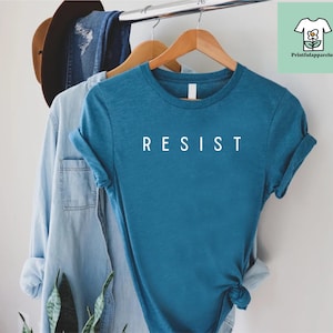 以下が含まれることがあります： 前面に白い文字で「RESIST」とプリントされたターコイズブルーのTシャツです。