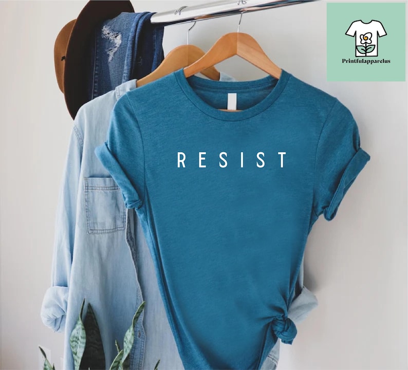 以下が含まれることがあります： 前面に白い文字で「RESIST」とプリントされたターコイズブルーのTシャツです。