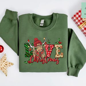 K&ouml;nnte beinhalten: Gr&uuml;ner Weihnachtspullover mit einem niedlichen Cartoon-Highland-Rind, das einen roten Weihnachtsmann-Hut tr&auml;gt und die Worte "Love Christmas".