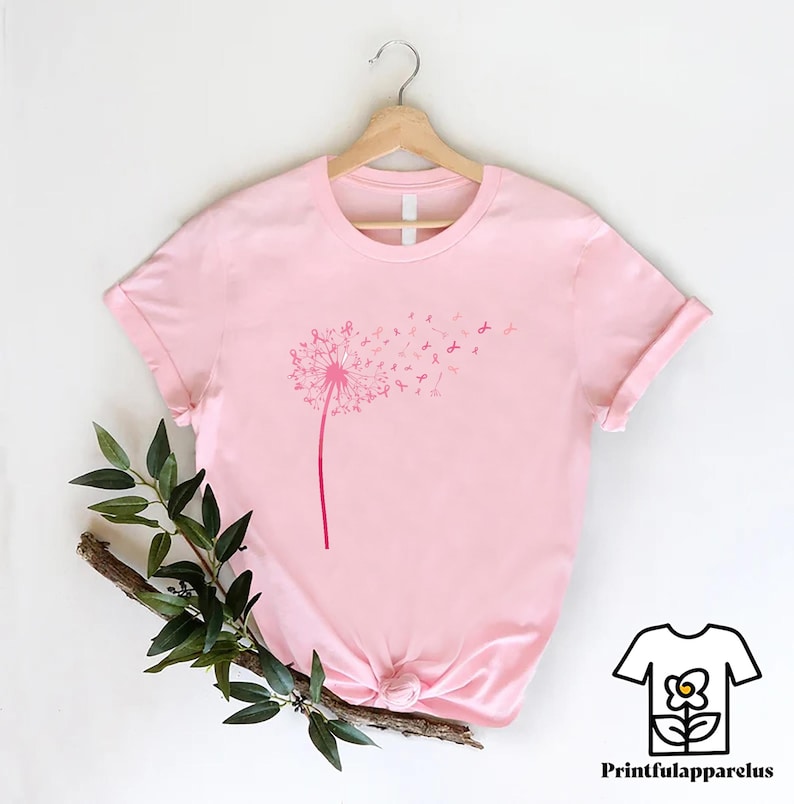 May include: A light pink t-shirt featuring a dandelion design with pink ribbons. The dandelion's stem and head are in a darker shade of pink. The shirt is displayed on a wooden hanger against a white background. The brand logo is in the bottom right corner.