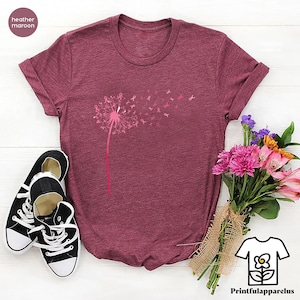 May include: Heather maroon t-shirt with a dandelion design, where the seeds are replaced with pink ribbons. The shirt is paired with black and white sneakers and a bouquet of pink and purple flowers. The text "Printfulapparelus" is visible.