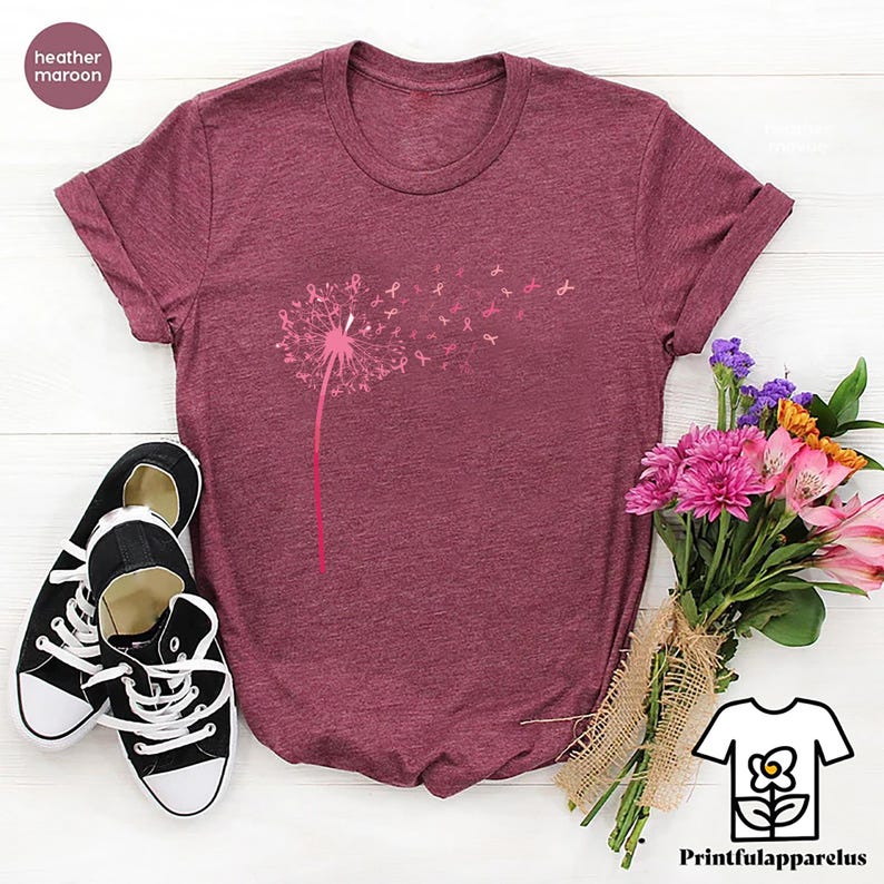 May include: Heather maroon t-shirt with a dandelion design, where the seeds are replaced with pink ribbons. The shirt is paired with black and white sneakers and a bouquet of pink and purple flowers. The text "Printfulapparelus" is visible.