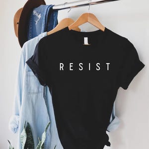 以下が含まれることがあります： 前面に白い文字で「RESIST」とプリントされた黒いTシャツ
