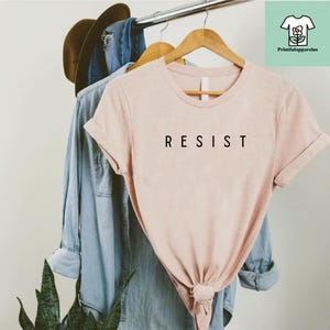 以下が含まれることがあります： 前面に黒い文字で「RESIST」とプリントされた薄いピンクのTシャツ。Tシャツは折り畳まれてハンガーにかけられています。