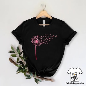 May include: Black t-shirt featuring a pink dandelion design with pink ribbon accents, symbolizing breast cancer awareness. The dandelion's seeds transform into pink ribbons, creating a visually striking and meaningful graphic. The shirt is displayed on a wooden hanger.