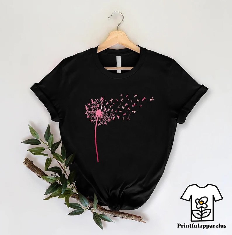 May include: Black t-shirt featuring a pink dandelion design with pink ribbon accents, symbolizing breast cancer awareness. The dandelion's seeds transform into pink ribbons, creating a visually striking and meaningful graphic. The shirt is displayed on a wooden hanger.