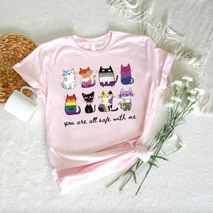 May include: A light pink t-shirt featuring a colorful illustration of nine cartoon cats, each with a unique design. The text "you are all safe with me" is printed below the cats. The shirt is made of soft material.