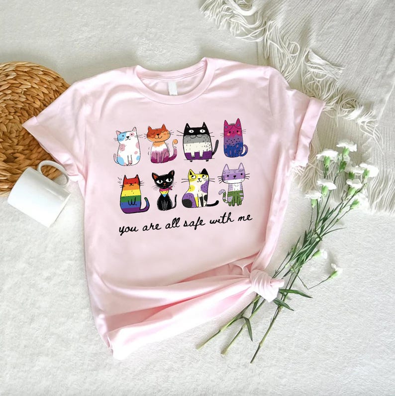 May include: A light pink t-shirt featuring a colorful illustration of nine cartoon cats, each with a unique design. The text "you are all safe with me" is printed below the cats. The shirt is made of soft material.