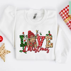 K&ouml;nnte beinhalten: Wei&szlig;er Pullover mit einem Weihnachtsmotiv, das eine schottische Hochlandkuh mit einem Weihnachtsmann-Hut und den Worten "Love Christmas" zeigt.