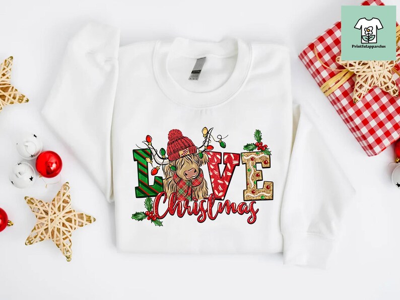 K&ouml;nnte beinhalten: Wei&szlig;er Pullover mit einem Weihnachtsmotiv, das eine schottische Hochlandkuh mit einem Weihnachtsmann-Hut und den Worten "Love Christmas" zeigt.