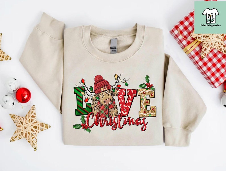 K&ouml;nnte beinhalten: Ein beiger Pullover mit einem weihnachtlichen Design, das eine schottische Hochlandkuh mit einem Weihnachtsmann-Hut und den Worten "Love Christmas" zeigt.