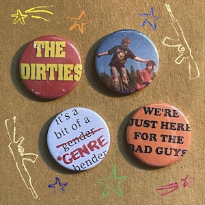 May include: Four circular pin badges with different designs and text. One reads "THE DIRTIES" in yellow on a red background. Another features a photo of two people. The third says "It's a bit of a gender *GENRE* bender". The last says "WE'RE JUST HERE FOR THE BAD GUYS".