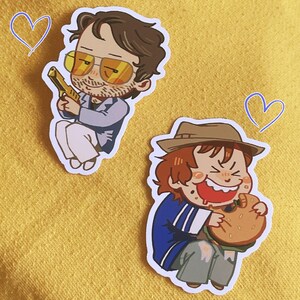 May include: Two die-cut stickers featuring cartoon characters. One shows a person with brown hair, yellow-tinted glasses, and a blue blazer holding a gold object. The other shows a person with a brown hat eating a large burger. Two blue heart outlines.