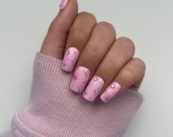 Peut inclure: Vernis à ongles rose et blanc avec de petites fleurs roses et des strass sur chaque ongle.