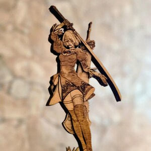 May include: A wooden figurine of a female character holding a sword. The figurine is standing on a base with grass-like details. The character is wearing a short dress and boots.