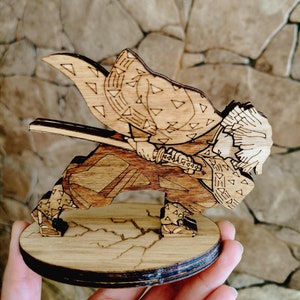 Może przedstawiać: Drewniana figurka postaci z anime Demon Slayer. Postać jest w pozie bojowej, trzymając miecz. Figurka jest szczegółowo rzeźbiona i pomalowana w odcieniach brązu.