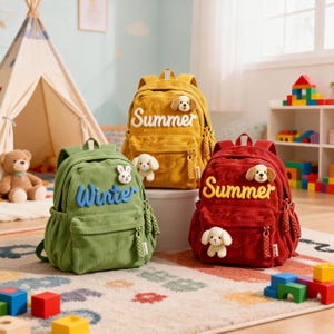 Pode incluir: Três mochilas de veludo cotelê em verde, amarelo e vermelho. Cada mochila tem a palavra "Winter" ou "Summer" bordada. As mochilas têm um pequeno animal de pelúcia preso.