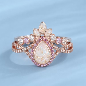Könnte beinhalten: Ein roségoldenes Ring-Set mit einem birnenförmigen Opal-Mittelstein, umgeben von rosa Edelsteinen. Der obere Ring hat ein Kronen-Design mit weißen Opal-Akzenten und kleinen, klaren Edelsteinen. Das Band hat ein verdrehtes Design mit kleinen, klaren Edelsteinen.