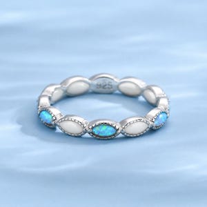 May include: A silver ring with a repeating pattern of oval-shaped blue opal stones and white enamel. The ring is set against a light blue background. The ring is marked with "925".