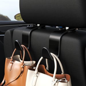 May include: Black car seat headrest hooks holding two handbags. One handbag is tan, the other is white. The hooks are made of black plastic and are designed to hang bags and other items in a vehicle.
