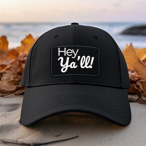 Può includere: Cappellino da baseball nero con una toppa bianca che dice "Hey Ya'll!" Il cappello è su una spiaggia sabbiosa con foglie autunnali.
