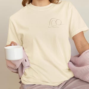 Op de afbeelding: Crèmekleurig T-shirt met een minimalistisch lijntekeningontwerp op de linkerborst. Het ontwerp toont twee verbonden vormen. De persoon houdt een witte mok vast en draagt een roze vest.