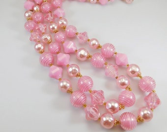 May include: A three-strand pink beaded necklace with various shapes and sizes of beads. The necklace features round, star, and faceted beads, with gold-tone metal accents and a clasp.