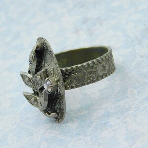 May include: A silver-toned ring with an ornate design. The ring features a central, textured oval shape with a small, clear gemstone and smaller accent stones. The band has a detailed pattern. The ring is set against a light blue background.