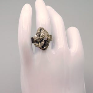 May include: A silver-toned ring with a leaf-like design. The ring features small, round embellishments and a central, iridescent stone. The ring is displayed on a white hand, showcasing its intricate details and vintage style.