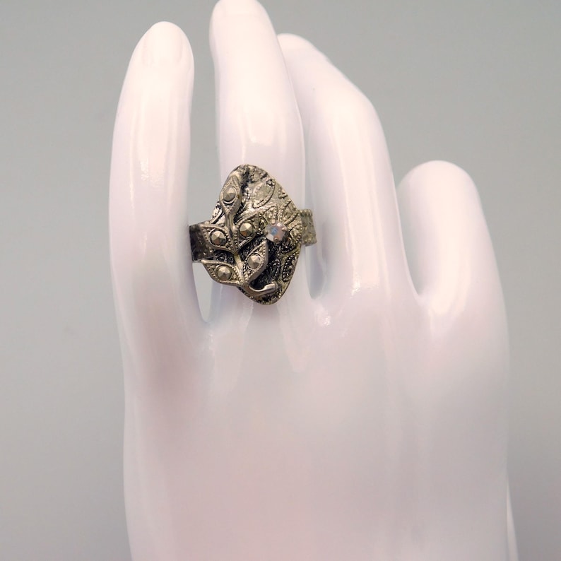 May include: A silver-toned ring with a leaf-like design. The ring features small, round embellishments and a central, iridescent stone. The ring is displayed on a white hand, showcasing its intricate details and vintage style.