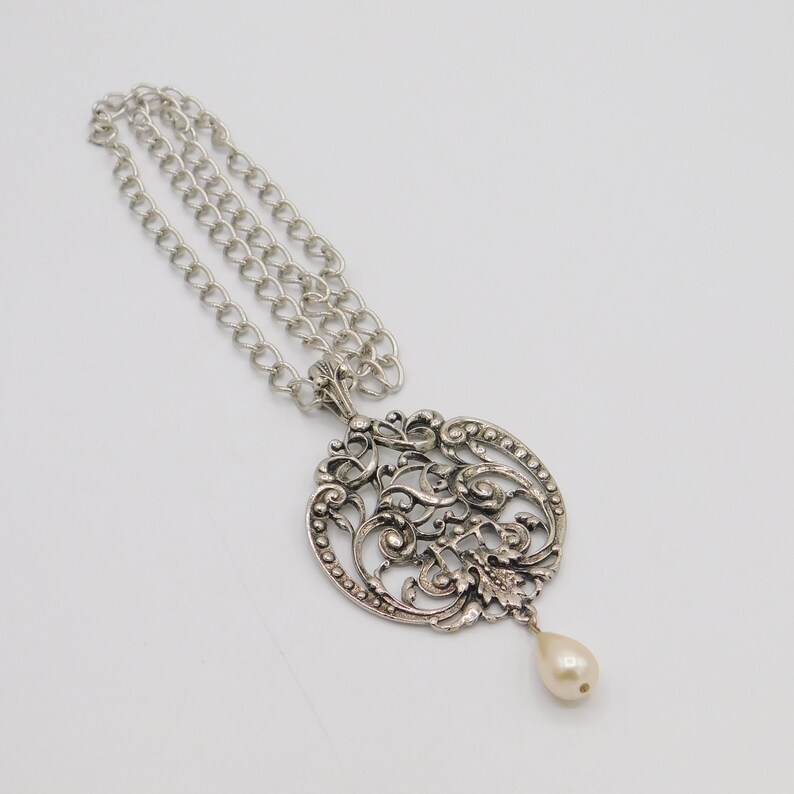 Puede incluir: Un collar de plata con un delicado colgante adornado con una perla. El colgante es de dise&ntilde;o circular con un intrincado trabajo de filigrana.