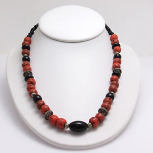 May include: A necklace with red and black beads, with silver-coloured metal spacers. The necklace is displayed on a white bust. The beads are of varying sizes and shapes, giving a handcrafted appearance. The necklace is a striking piece of jewellery.