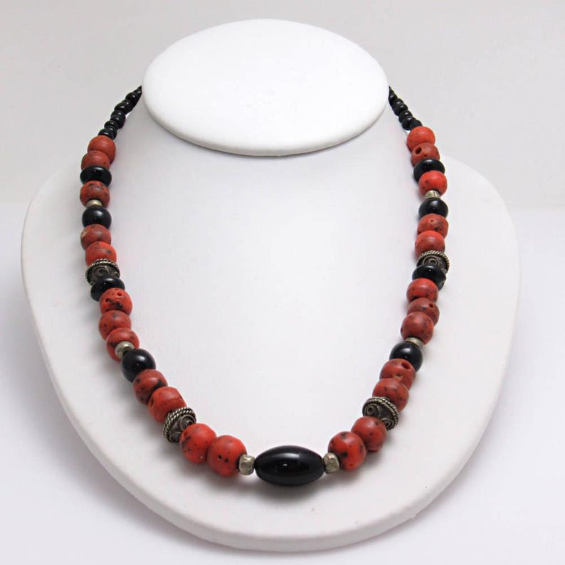 May include: A necklace with red and black beads, with silver-coloured metal spacers. The necklace is displayed on a white bust. The beads are of varying sizes and shapes, giving a handcrafted appearance. The necklace is a striking piece of jewellery.