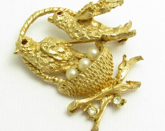Op de afbeelding: Gouden broche met twee vogels die op een tak zitten met een nest met drie nep parel-eieren. Een vogel heeft rode strass-ogen.