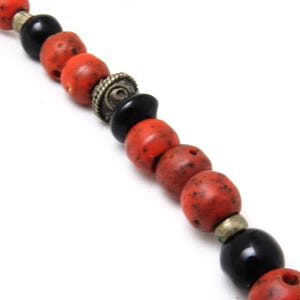 May include: A close-up of a necklace with red, speckled beads and black beads. The necklace also features silver-toned metal accents. The beads are strung together, creating a unique design. The necklace is set against a white background.